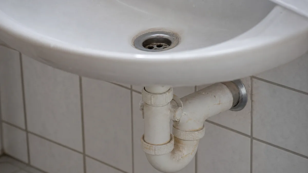 Detailaufnahme eines Waschbecken-Abflusses mit sichtbarem Siphon darunter in einem hellen Badezimmer